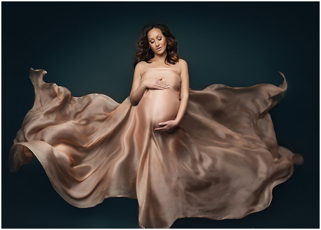 Pregnant woman with flowing champagne silk fabric on a dark teal backdrop, luxury maternity portrait